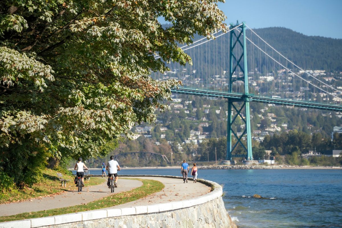 Two cyclists riding on a path in Stanley Park with the Lions Gate Bridge in the background.